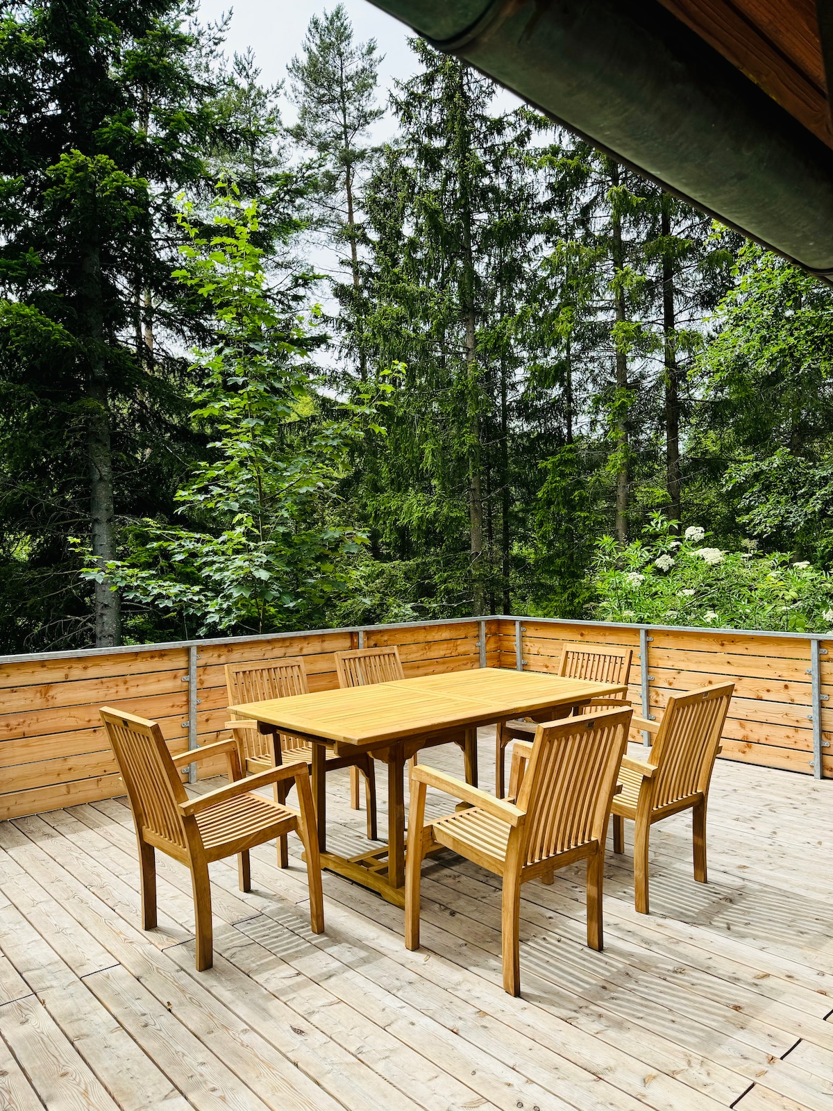A wooden terrace features a six-seat outdoor dining set surrounded by lush greenery. The natural wood finish complements the serene forest backdrop, providing a peaceful spot for meals. The terrace is bordered by timber railings, enhancing the connection with nature.