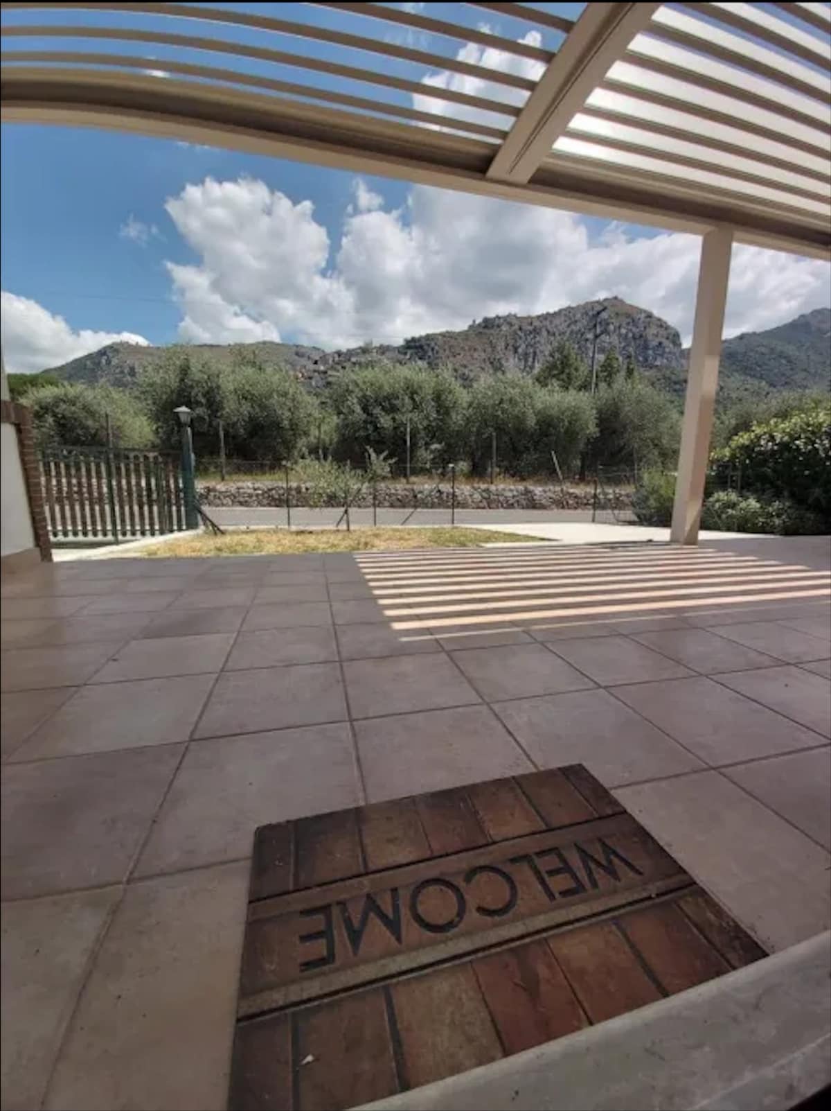 A welcoming outdoor area features a tiled floor under a shaded patio. The landscape includes a view of mountains and greenery beyond the fence, with clouds scattered in the sky, creating a serene atmosphere.