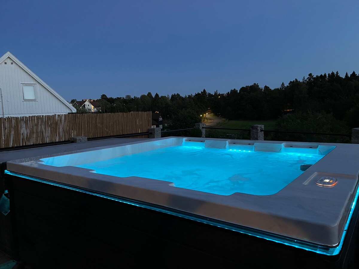 A hot tub is illuminated in soft blue light, set against a darkening twilight sky. The surrounding area features a natural landscape with trees and distant houses, providing a serene backdrop for relaxation.