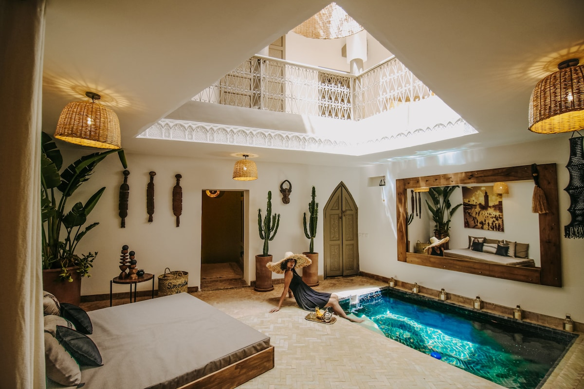 An indoor pool is framed by wooden planks, reflecting natural light from the skylight above. Surrounding the pool, potted plants add a touch of greenery, while a large mirror enhances the sense of space. Traditional Moroccan decor elements are displayed on the walls.