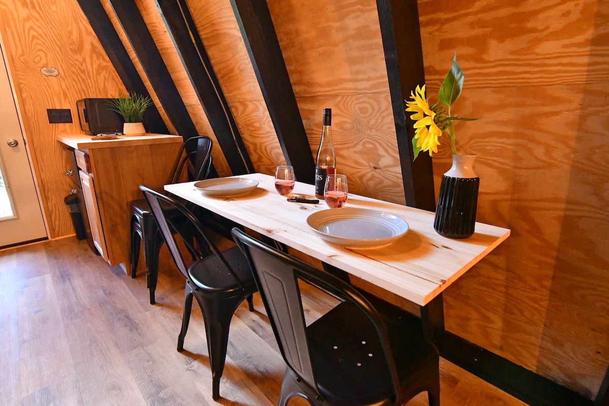 A dining area features a wooden table set for two, with plates and glasses positioned atop it. Black metal chairs accompany the table, and a kitchen counter is visible in the background. A sunflower in a vase adds a touch of color against the warm wood paneling.