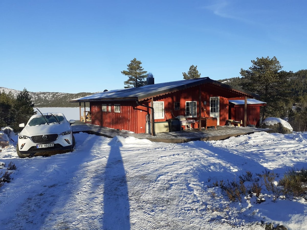 A red cabin is set against a snowy landscape, featuring large windows that provide views of the surrounding nature. A car is parked nearby on a snowy driveway. Trees and mountains are visible in the background, enhancing the serene atmosphere.