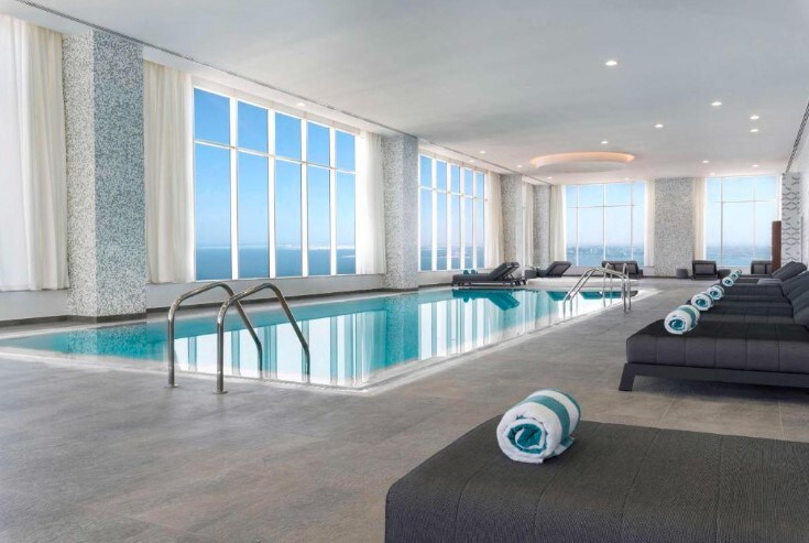 An indoor swimming pool is highlighted by large windows that allow natural light to enhance the space. Lounge chairs with rolled towels line the poolside, and a serene view of the water can be seen through the expansive glass panels.