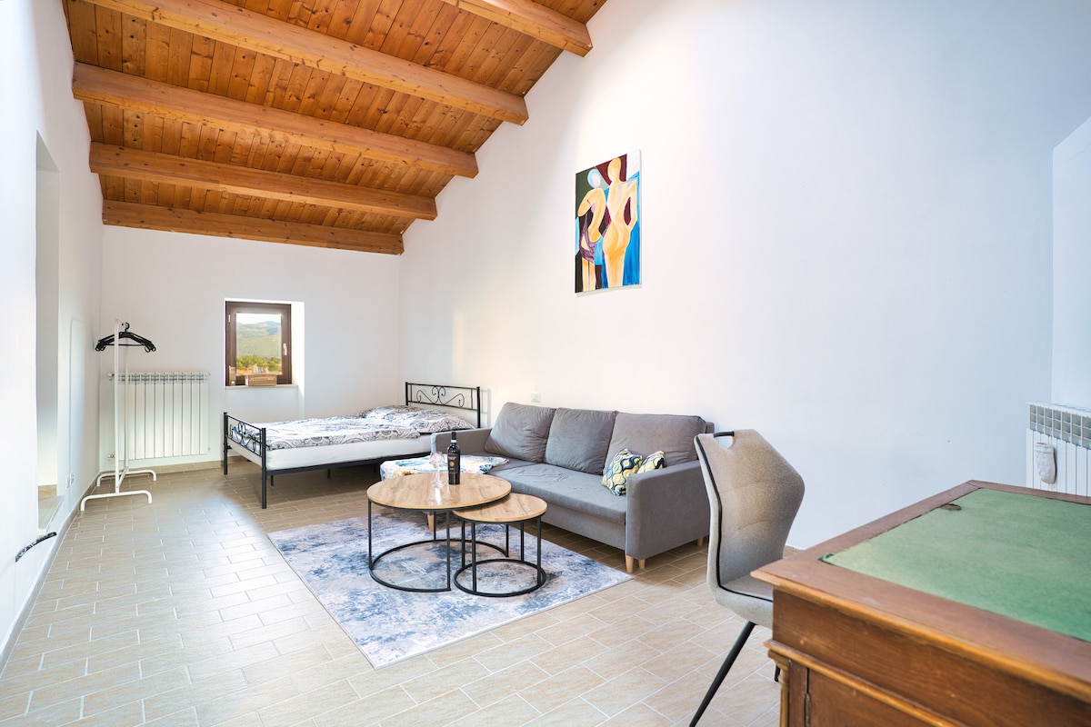 A spacious room features wooden beams across the ceiling, with a large window allowing natural light. A cozy bed and a comfortable sofa are positioned near a coffee table. A piece of art adds a touch of color to the minimalist decor.