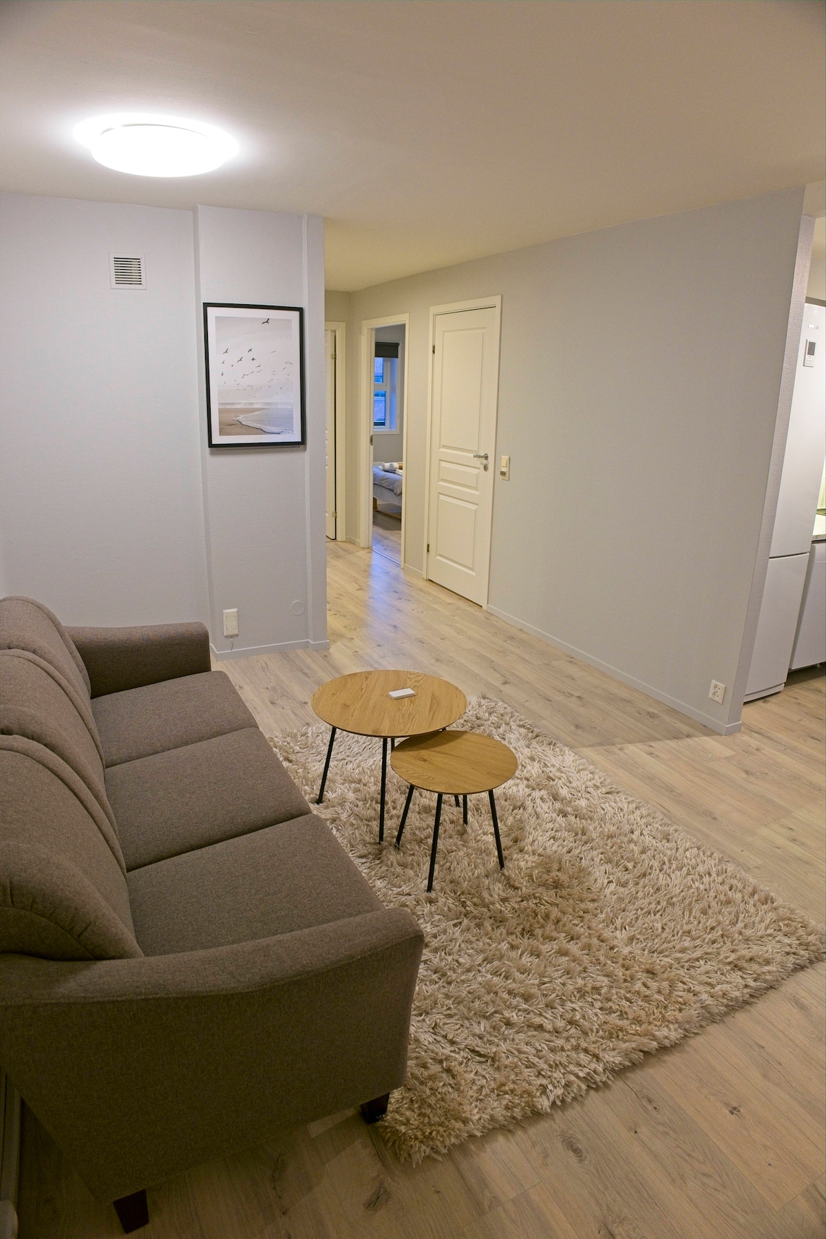 A modern living area is portrayed, featuring a comfortable gray sofa facing two small wooden tables placed on a soft rug. Light wood floors and neutral wall colors create a spacious feel, with a doorway visible leading to another room.