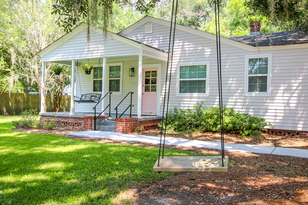 A charming exterior view of a light-colored home is depicted, featuring a welcoming front porch with a simple swing hanging from a sturdy branch above. Lush green grass surrounds the house, complemented by well-maintained landscaping and tall trees in the background.