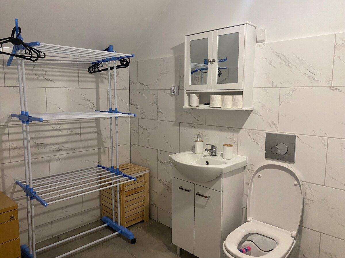A functional bathroom space includes a modern toilet and a compact sink with a cabinet above. A drying rack stands in one corner, providing ample hanging space. Neutral tiled walls enhance the room's clean appearance, complemented by a wooden storage unit nearby.