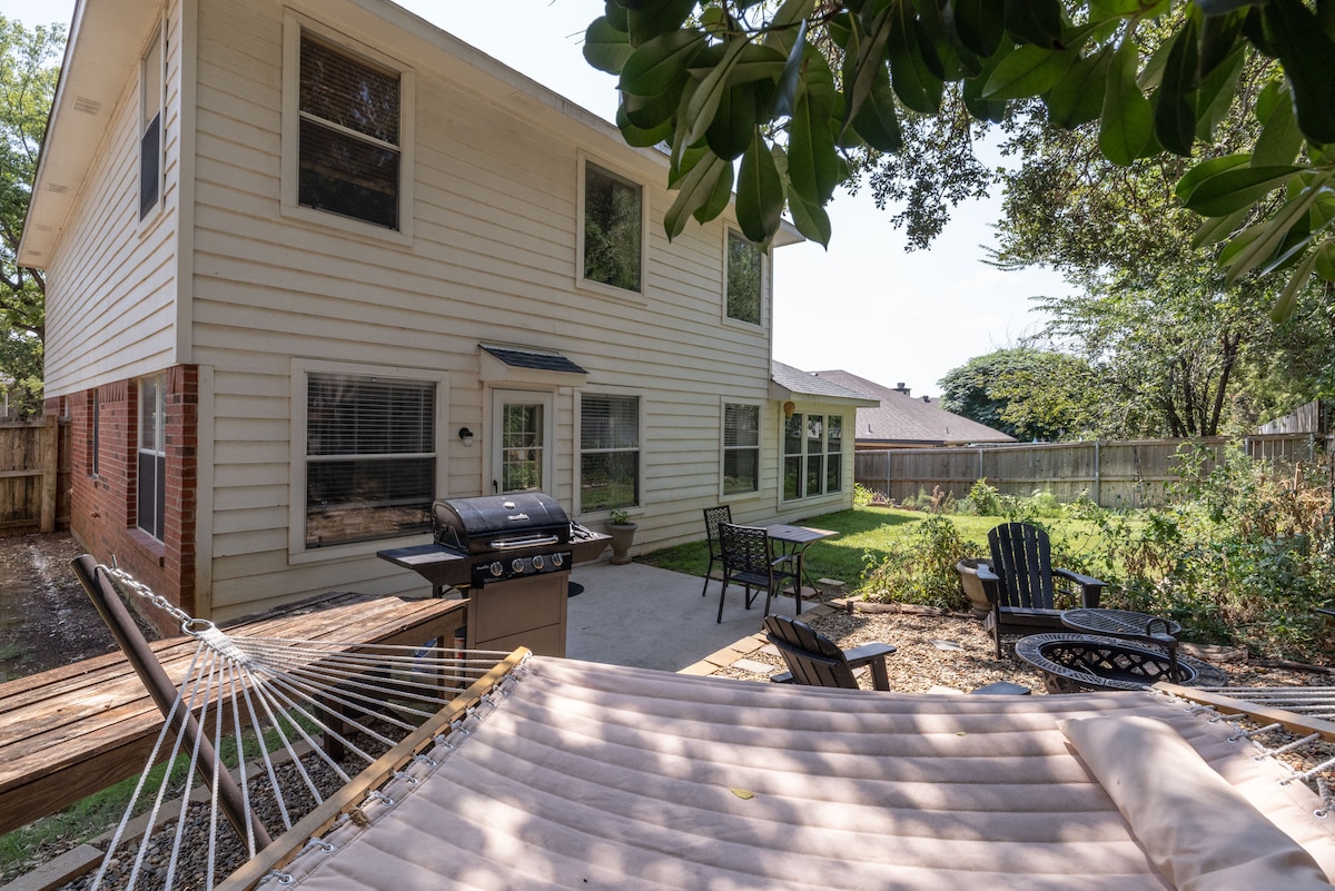 A spacious backyard is shown with a well-maintained lawn. A hammock is positioned in the foreground, while a grill and seating area with chairs are viewed behind it. The two-story home with multiple windows provides a light and airy atmosphere.