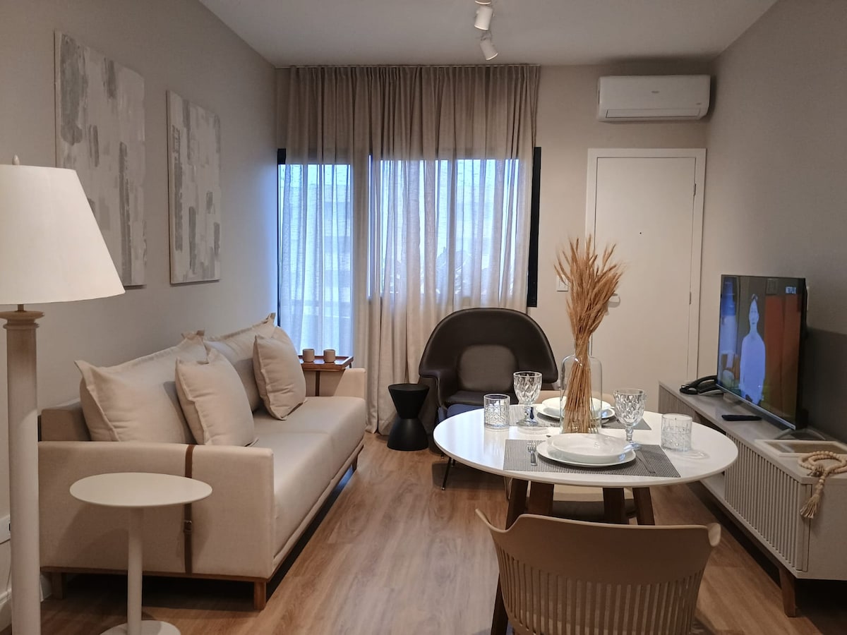 A well-arranged living area showcases a light-colored sofa accompanied by a round dining table set for four. Natural light enters through large windows, complemented by sheer curtains. A smart TV is positioned in view, and decorative elements include a floor lamp and a minimalist chair.