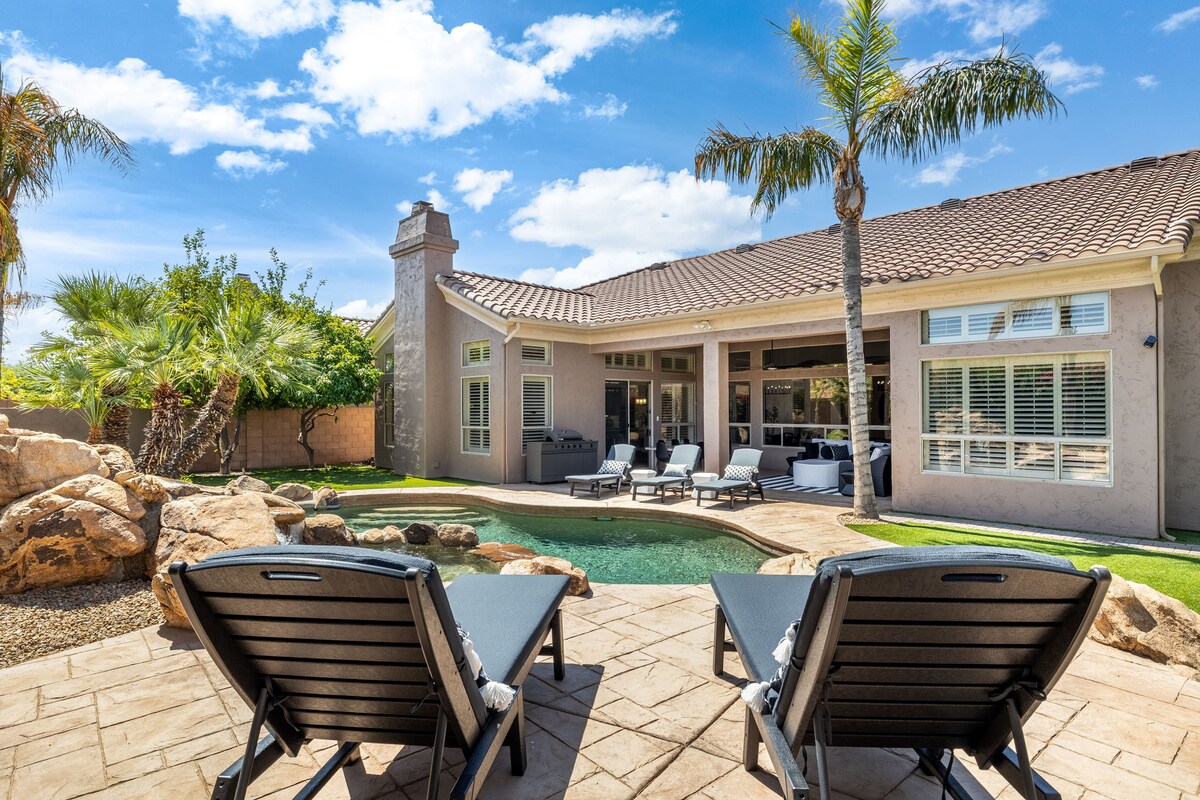 A serene backyard features a rectangular heated pool surrounded by natural stone and tropical landscaping. Two lounge chairs are positioned by the pool's edge, under the clear blue sky with scattered clouds. The spacious home can be seen in the background, boasting large windows and a covered patio.