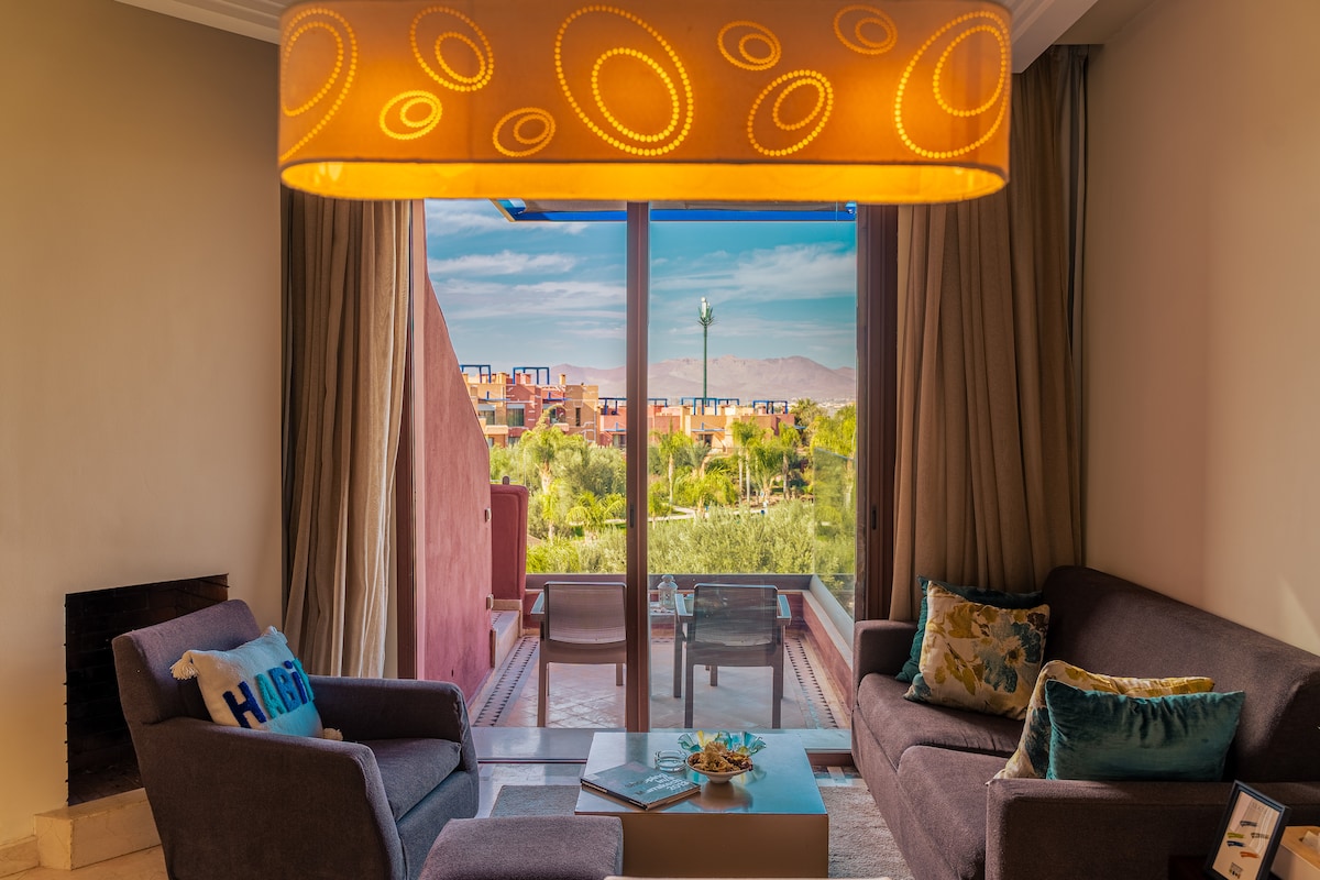 A comfortable living area is presented, featuring two armchairs and a sofa arranged around a low coffee table. A large window offers a view of the lush greenery and distant mountains, while soft lighting from a circular lamp enhances the space.