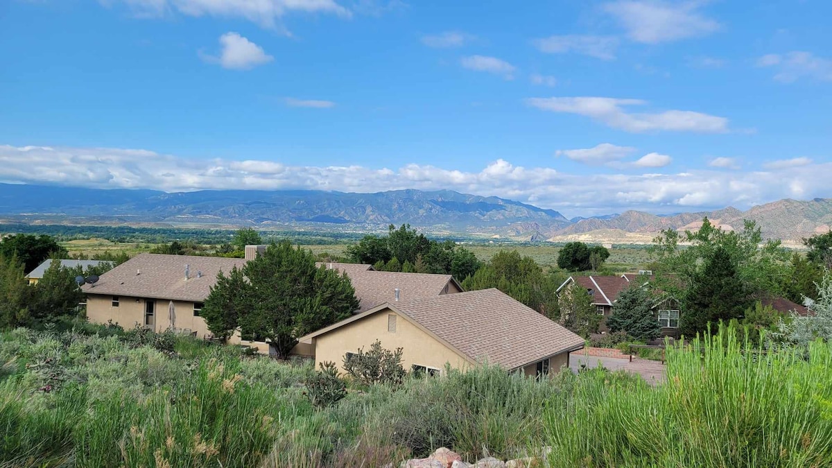 A scenic view features the vacation home nestled among greenery, showcasing a backdrop of rolling mountains and a blue sky with scattered clouds. Natural vegetation and shrubs fill the foreground, while neighboring homes can be seen in the distance.