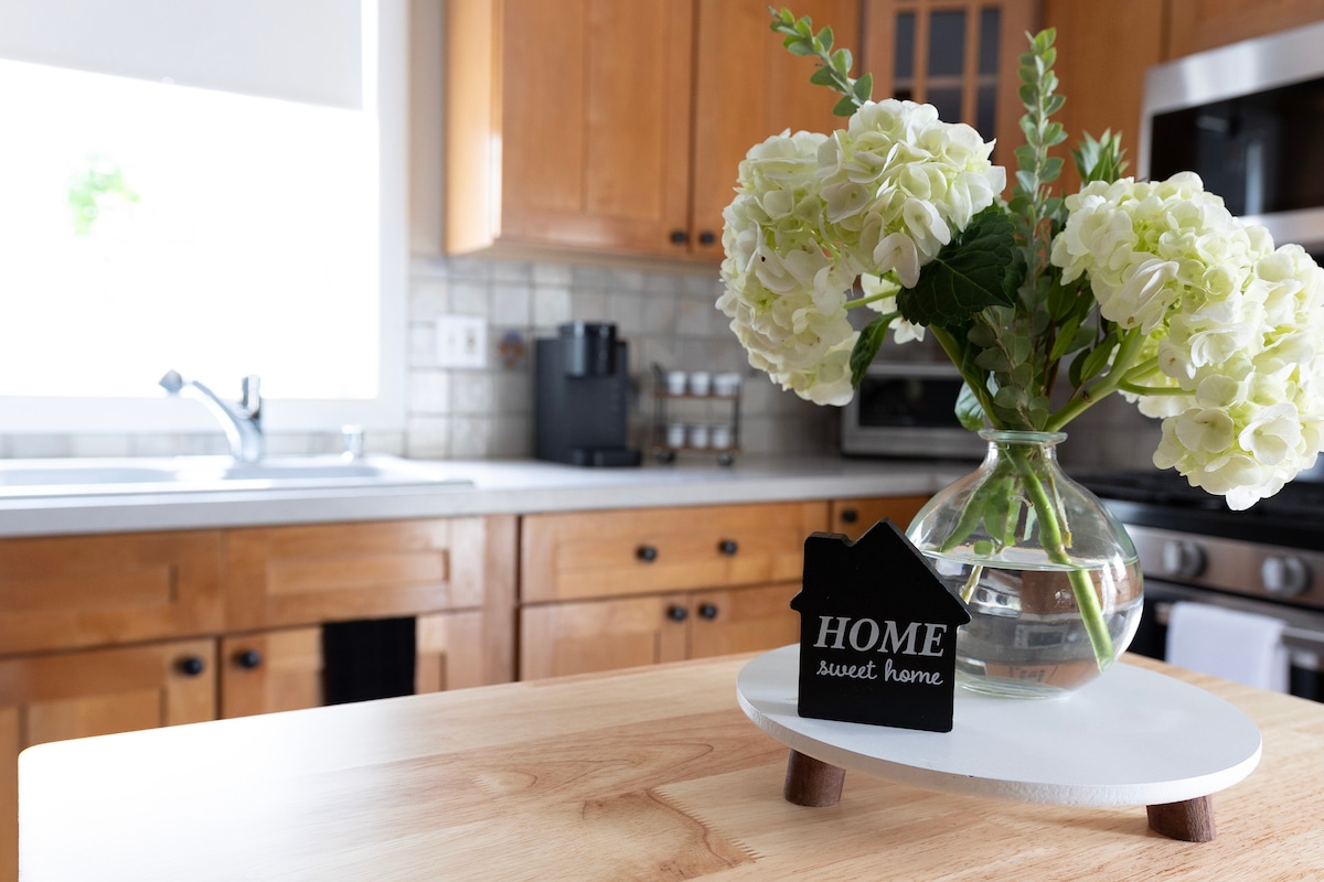 A modern kitchen is depicted, featuring polished wooden cabinets and stainless steel appliances. A glass vase holds white hydrangeas, accompanied by a decorative sign that reads 'HOME sweet home.' Natural light enters through the window, illuminating the warm tones of the space.