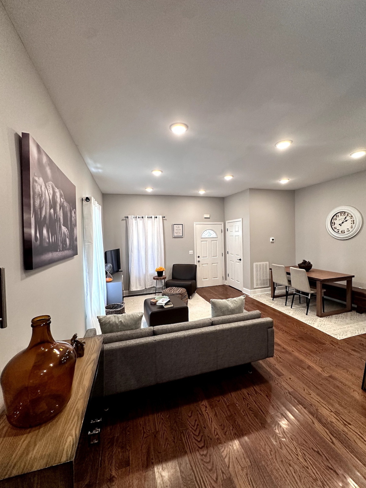 A modern living area is presented with a neutral color palette, featuring a comfortable sofa and a stylish seating arrangement. Natural light enters through sheer curtains, illuminating the space. A dining table sits adjacent, with a wall clock adding a welcoming touch.