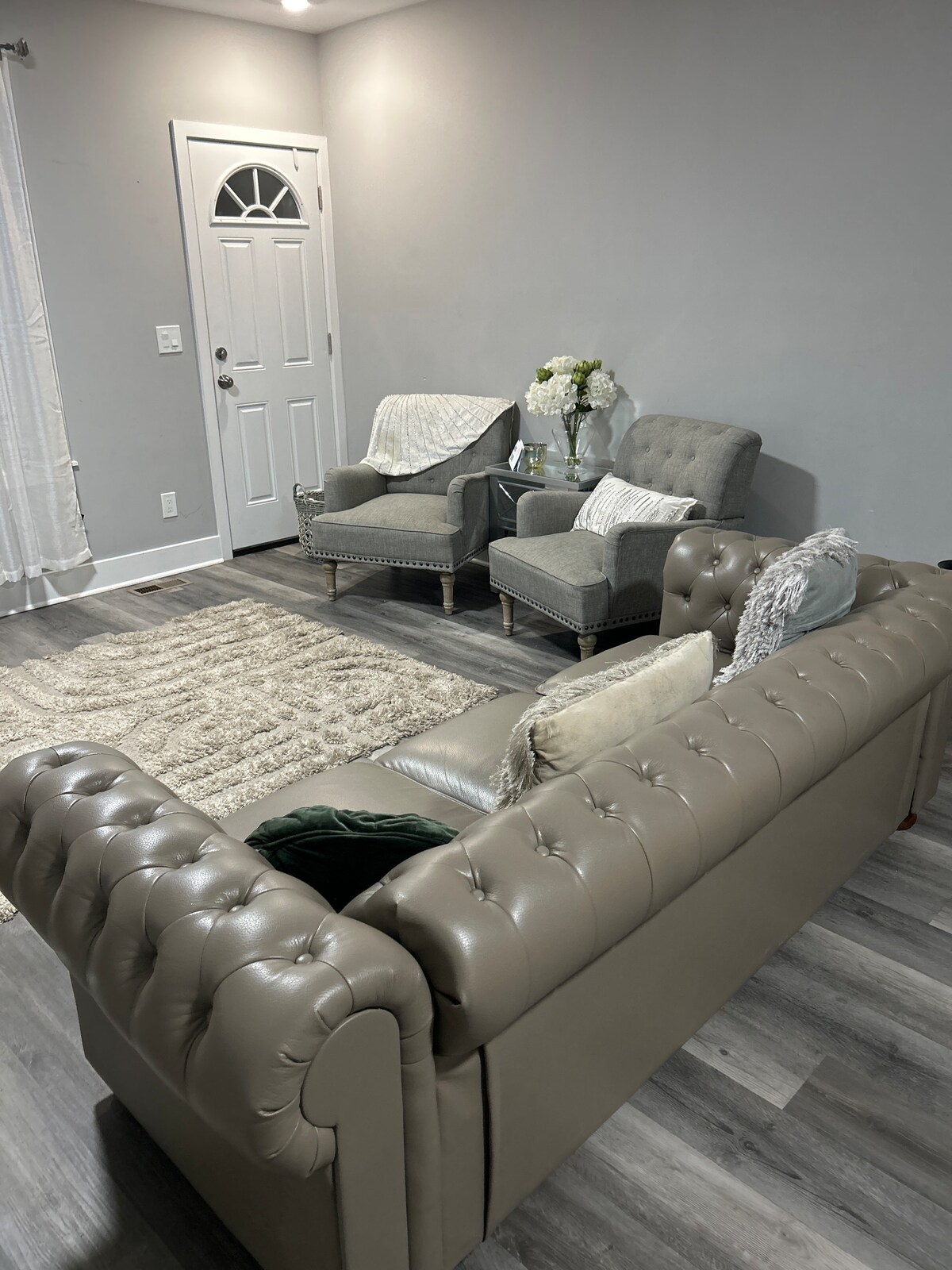 A relaxed seating area showcases a tufted beige sofa facing two light gray armchairs with soft throw blankets draped over them. A plush area rug is laid beneath, while a small table holds a vase with fresh flowers. Natural light filters through a nearby window.