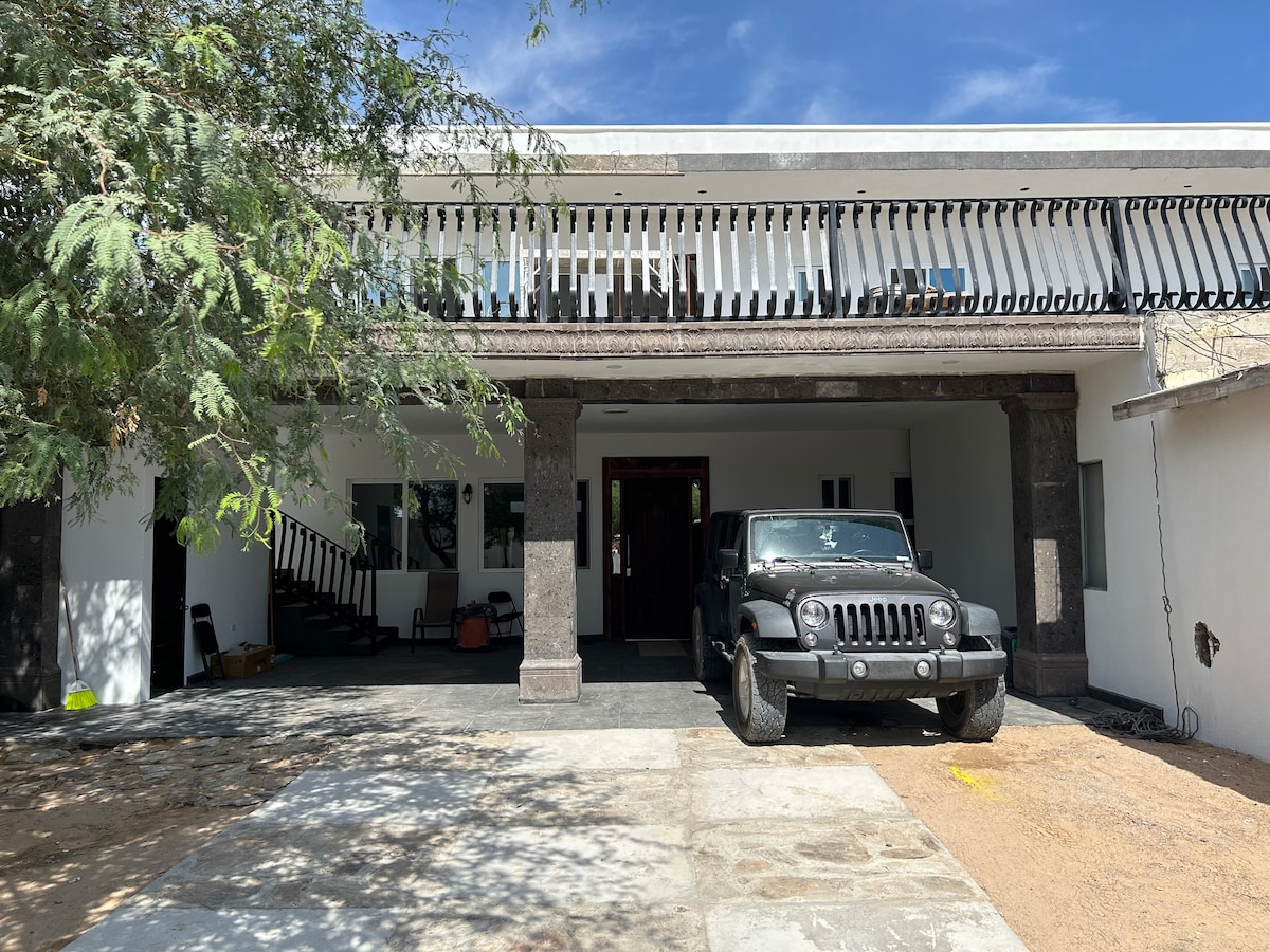 The entrance features a spacious outdoor area with a concrete pathway leading to a double-door entry. A black SUV is parked beside the path, surrounded by decorative foliage. The two-story structure showcases an upper balcony with railings, inviting natural light into the space.