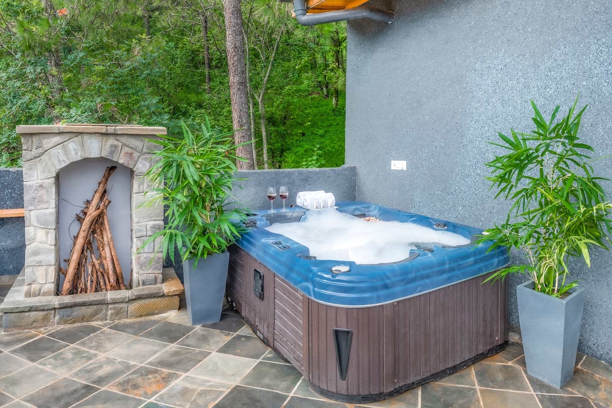 A private outdoor jacuzzi is surrounded by lush greenery, providing a tranquil retreat. Stone walls and potted plants frame the area, while a cozy fireplace made of natural stone adds warmth. Two glasses are placed on the edge of the jacuzzi, inviting relaxation.