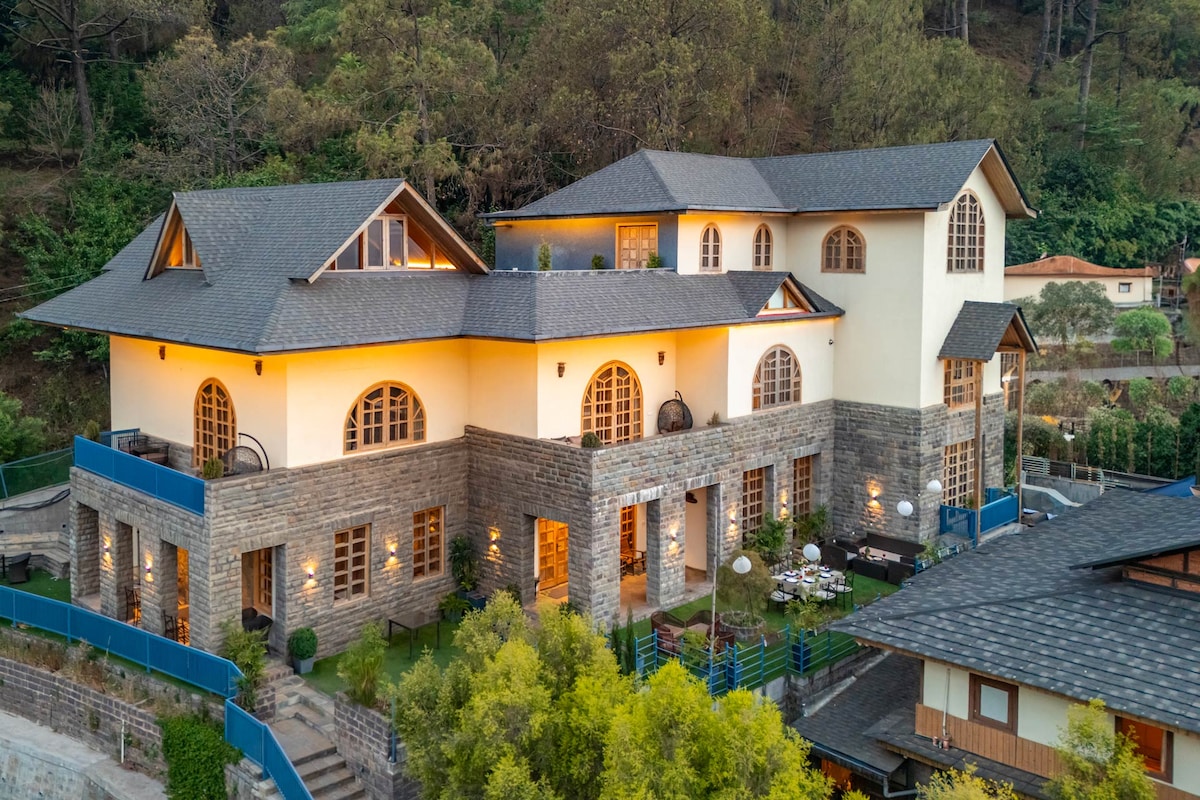 An aerial view showcases the striking architecture of the villa, featuring a blend of stone and modern design elements. The structure is illuminated softly in the evening, surrounded by lush greenery and thoughtful landscaping, including seating areas visible in the outdoor spaces.