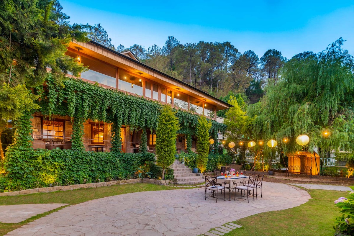 The villa is enveloped in lush greenery, featuring a stone patio adjacent to a well-kept lawn. Warm outdoor lighting enhances the ambiance, and a dining set is positioned to accommodate guests. Surrounding trees provide a natural backdrop, contributing to a serene environment.