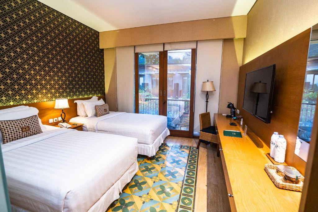 This guestroom features two spacious beds covered in crisp white linens, positioned against a backdrop of textured wallpaper. A sliding glass door opens to a balcony with garden views. A wooden desk with a lamp and a flat-screen TV are also visible.