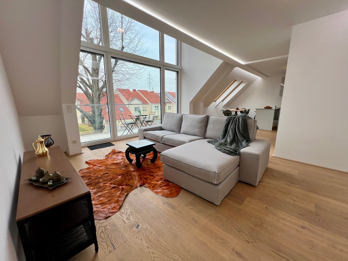 A modern living area features a spacious gray sofa with a decorative throw draped over it. A rustic coffee table sits on a textured area rug. Large windows provide abundant natural light, with views of neighboring rooftops and trees visible outside.