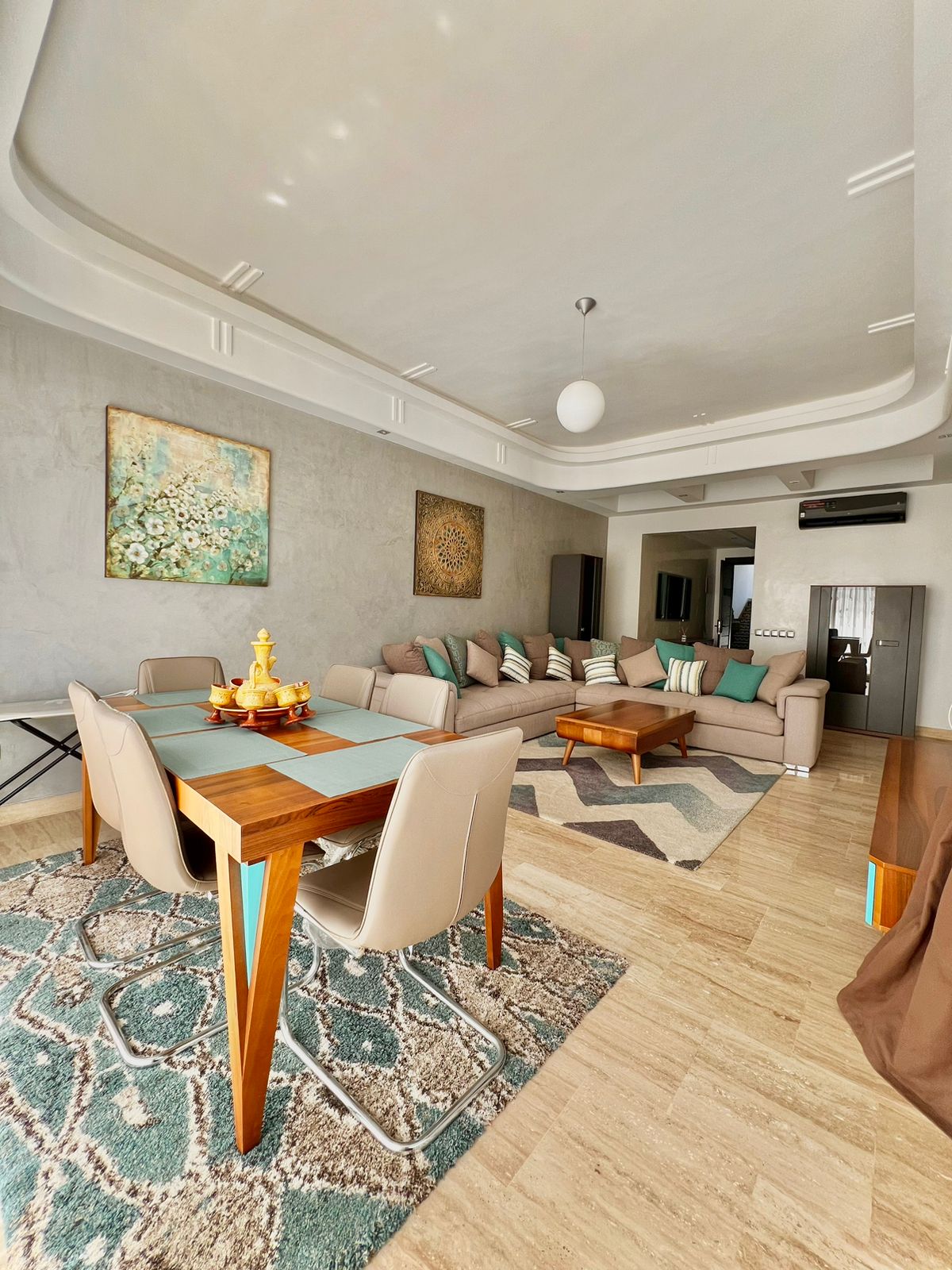 A spacious living area is furnished with a large, comfortable sofa accented by colorful cushions. A dining table with light-colored chairs is set against a textured wall. Natural light floods the room, highlighting the warm tones of the decor and the stylish area rug.