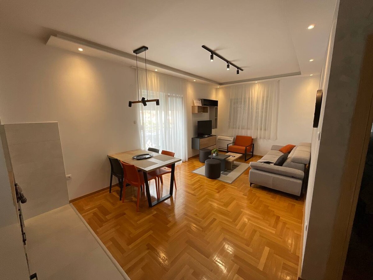 A modern living area is presented with hardwood flooring and natural light filtering through sheer curtains. A light-colored sofa complements an orange accent chair, while a stylish dining table is surrounded by four chairs. A sleek television stand enhances the room's aesthetic.