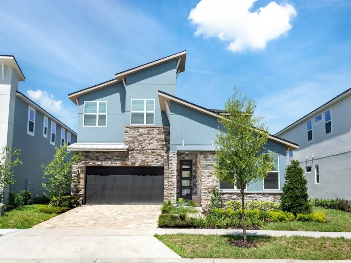 A modern two-story home features a stone-accented facade and a well-maintained garden. The driveway leads to a two-car garage, while large windows offer views of the surrounding landscape. The roofline adds visual interest, and the sky is bright with scattered clouds.