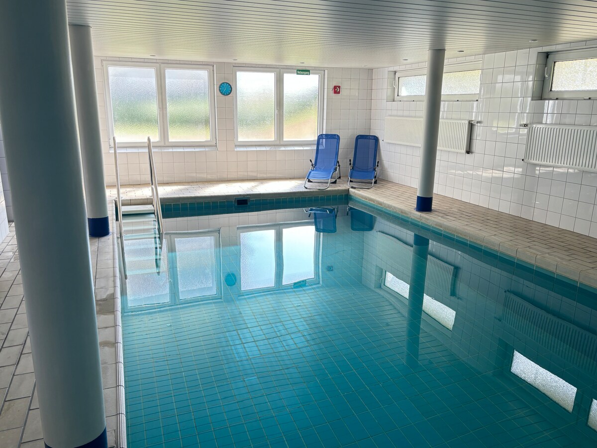 An indoor pool area is shown, featuring pristine blue water reflecting the light from large windows. Two blue lounge chairs sit beside the water, positioned against white tiled walls and columns. The space appears bright and inviting, ideal for relaxation.
