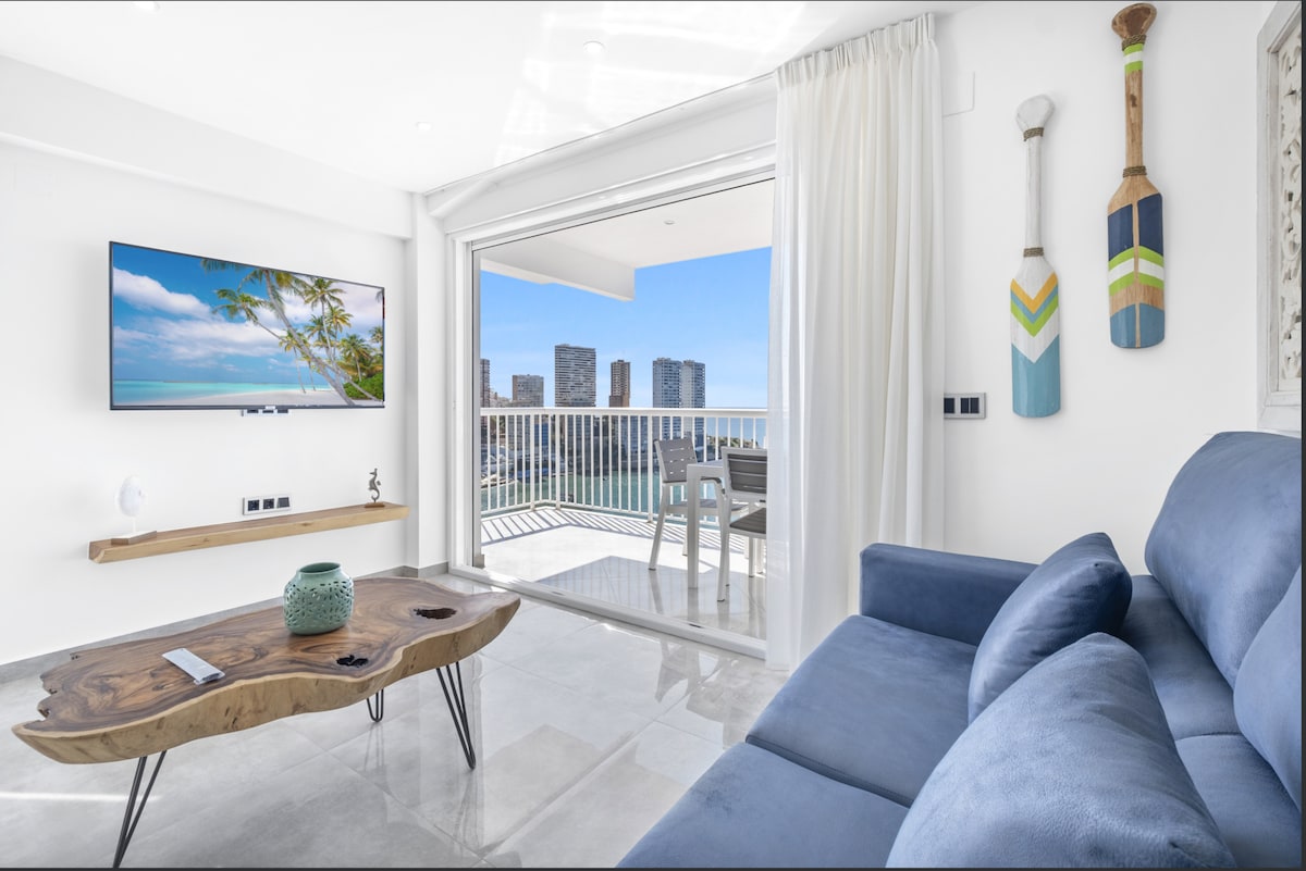 A light-filled living area is showcased with a comfortable blue sofa and a wooden coffee table. Large glass doors open to a balcony, offering panoramic views of the sea and city. A television is mounted on the wall, and decorative paddle accents are visible.