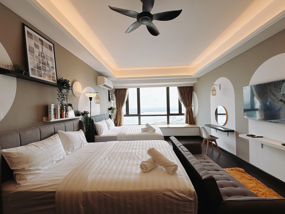 The spacious studio apartment features two queen-sized beds with fresh linens, situated against warm-toned walls. A modern ceiling fan hangs above, and large windows provide a full sea view. A cozy sofa and a wall-mounted television complete the inviting setting.