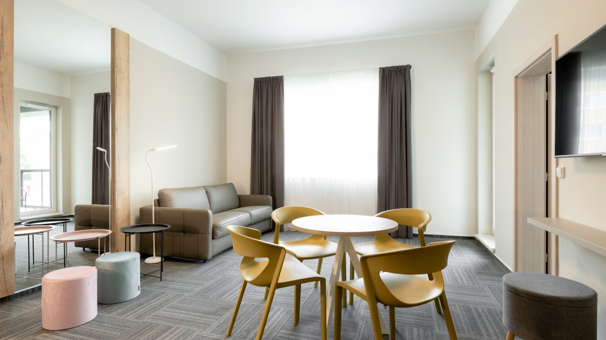 A bright living space is furnished with a comfortable sofa and a round dining table surrounded by four chairs. A large window allows natural light to fill the room, while soft curtains frame the view. Various small tables, a floor lamp, and stools are also present.