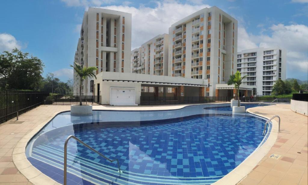 A large swimming pool is featured in the foreground, showcasing a mosaic tile pattern in shades of blue. Surrounding buildings, characterized by modern architecture, loom in the background amidst landscaped greenery. The area is designed for relaxation, with ample space for lounging.