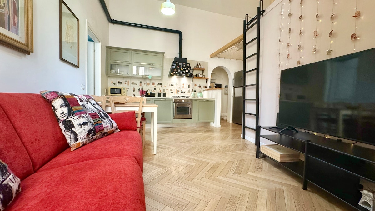 A living area is displayed with a red sofa adorned with decorative pillows. A sleek black television is positioned on a wooden stand. The open kitchen features a unique modern design with gray cabinetry and an island, while light wooden flooring extends throughout the space.