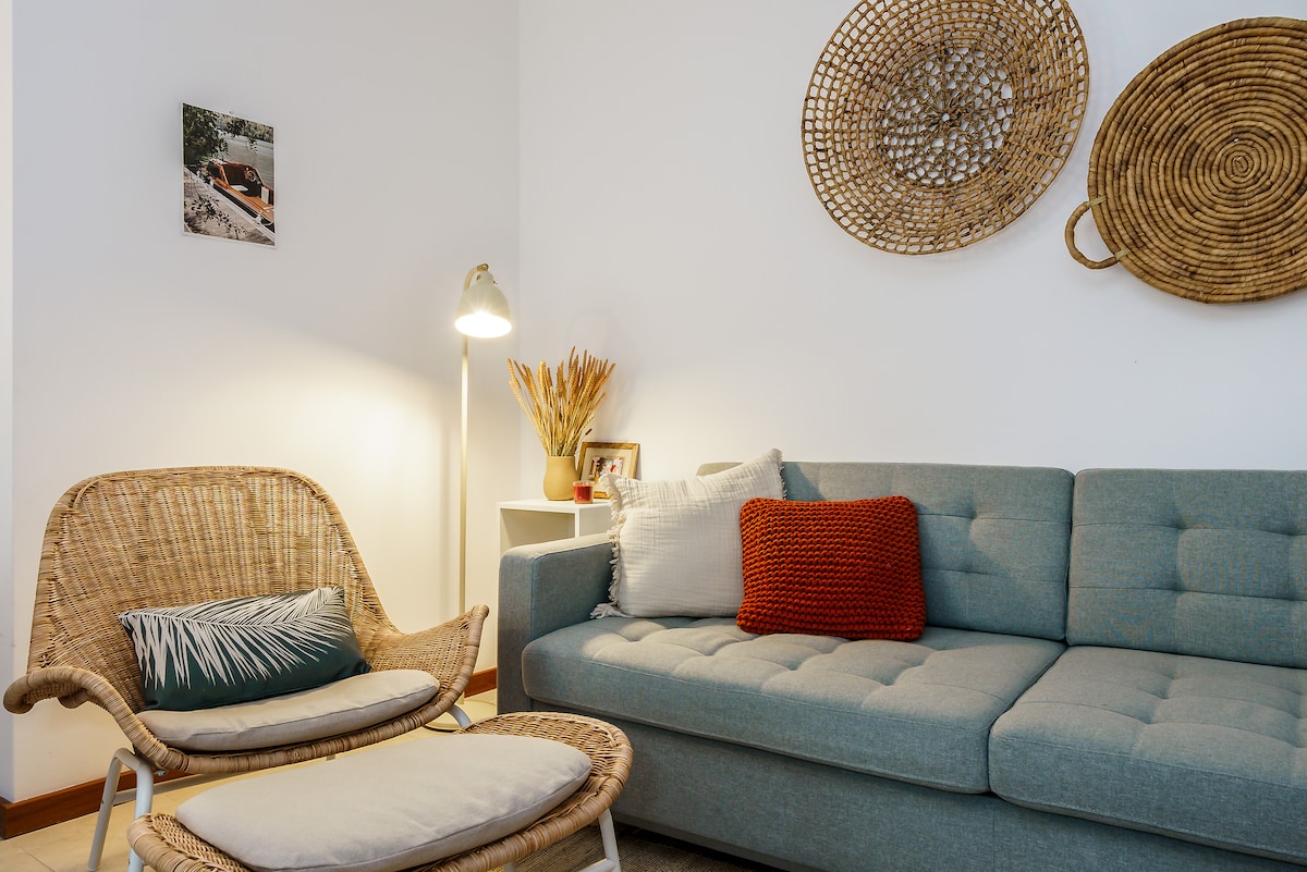 A cozy living area features a gray upholstered sofa adorned with textured pillows. A rattan chair and matching footrest provide additional seating. The space is illuminated by a floor lamp, and wall decor includes woven baskets, enhancing the natural aesthetic.