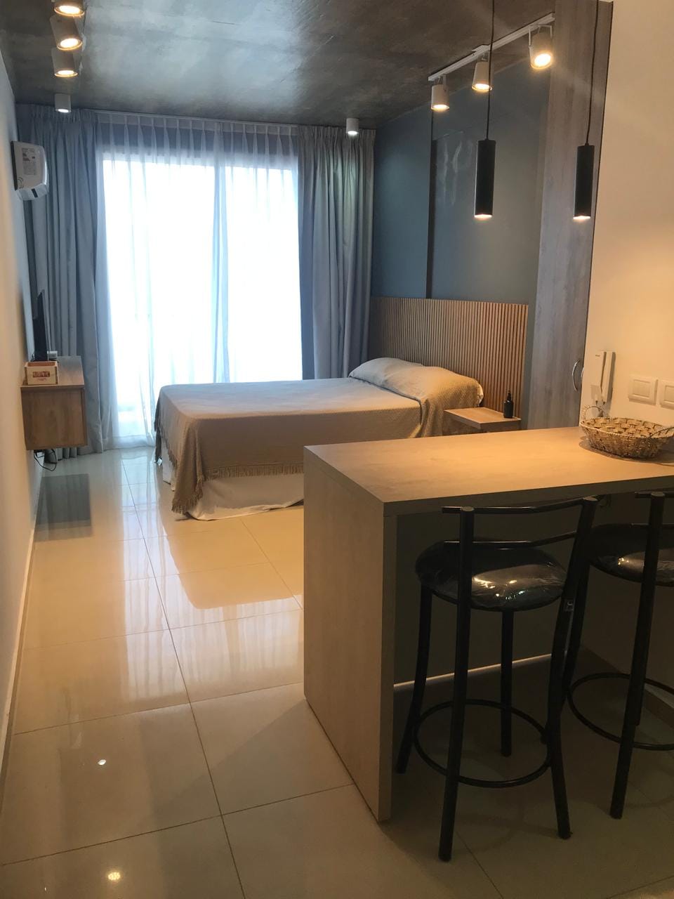 An inviting studio space showcases a spacious bed dressed in neutral linens, complemented by warm wood accents. A small dining area with two stools is positioned near a well-equipped kitchen. Large windows allow natural light to fill the room, enhancing its modern atmosphere.
