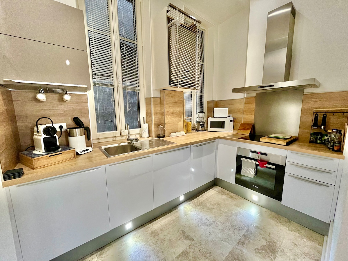 A modern kitchen is presented with sleek, glossy cabinetry and a minimalist design. Stainless steel appliances, including an oven and microwave, are clearly visible. The countertop features essential kitchen tools and has good natural light from tall windows with blinds. Soft lighting under the cabinets enhances the workspace.