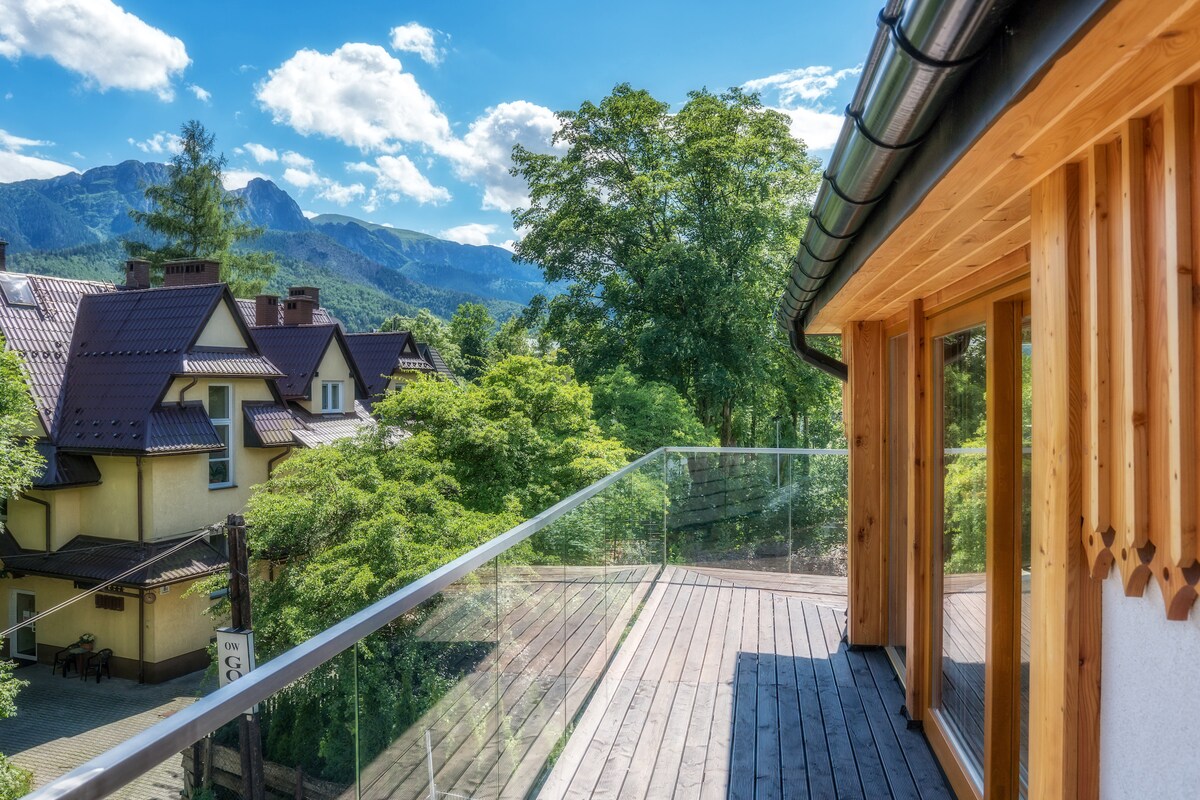 A spacious balcony features wooden planks and transparent railings, offering expansive views of lush greenery and distant mountains. Nearby houses are visible, nestled amongst trees, contributing to a serene outdoor atmosphere.