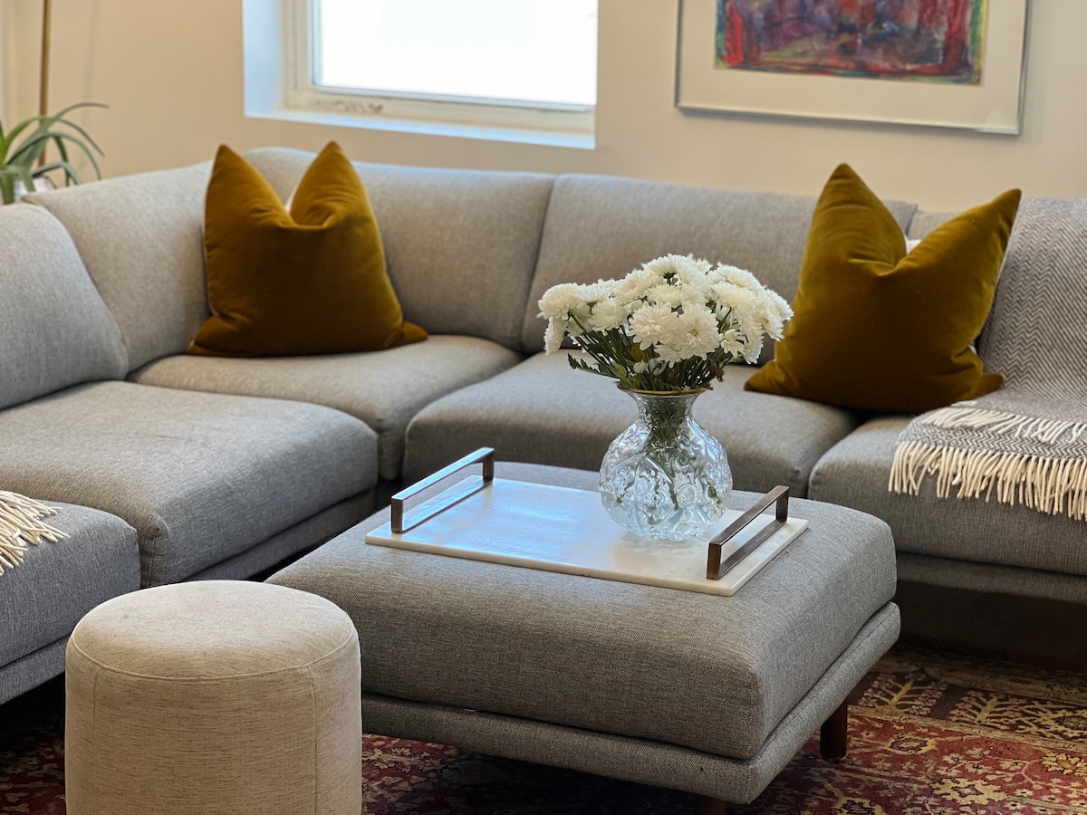 A modern sectional sofa with soft gray upholstery features two decorative yellow pillows. A clear glass vase filled with white flowers sits atop a low coffee table, which is styled with a wooden tray. A round pouf in a neutral tone is positioned nearby.