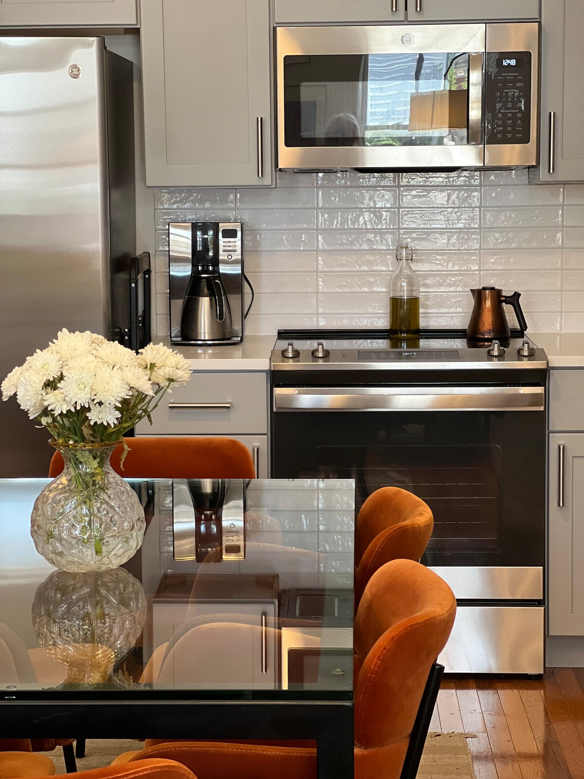 A modern kitchen space is showcased, featuring stainless steel appliances, including a microwave and stove. A glass dining table with orange velvet chairs is positioned in the foreground, accompanied by a floral arrangement in a clear vase. Light reflects off the smooth surfaces, creating a bright atmosphere.