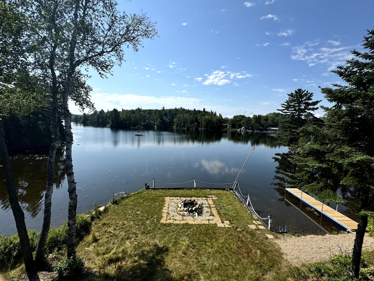 A serene lakeside view features a grassy area surrounding a stone fire pit, bordered by trees. The calm water reflects the clear blue sky, and a wooden dock extends toward the lake. A sandy shoreline invites relaxation and water activities.
