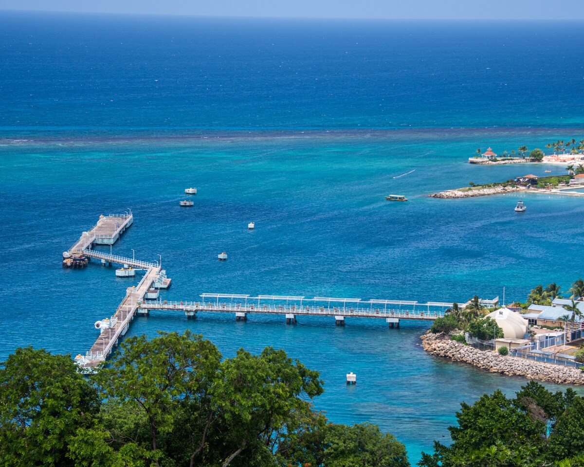 A panoramic view captures a serene harbor with a long wooden pier extending into the blue waters. Small boats drift nearby, while the shoreline is lined with lush greenery and buildings, all under a clear sky.