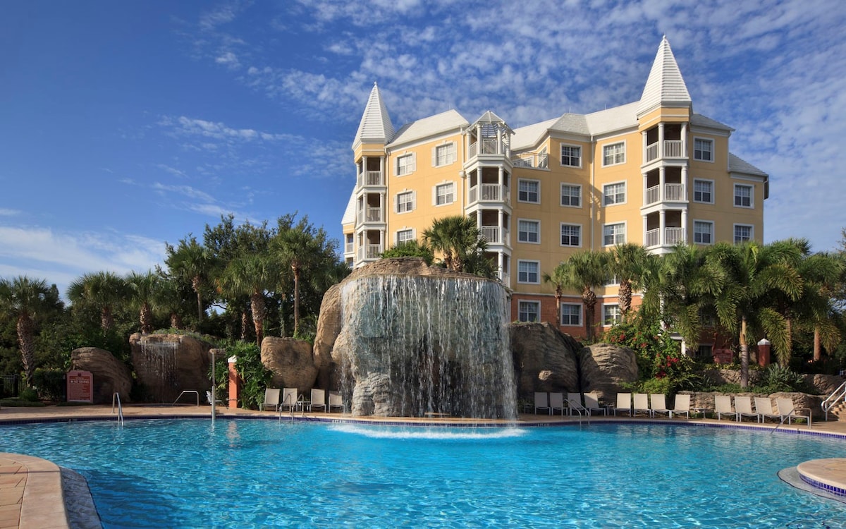 A large, inviting pool is surrounded by lush palm trees and vibrant landscaping. A waterfall cascades over a rocky feature, creating a natural focal point. The pastel yellow building with pointed roofs adds to the tropical ambiance of the space.