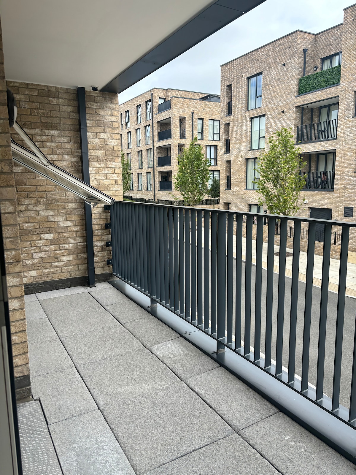 The private balcony features a sleek railing and is accessed through sliding doors. A spacious area is provided with grey paving stones, offering a view of the surrounding brick buildings and green trees, creating a serene outdoor space.