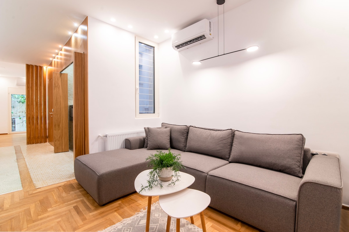A spacious lounge area is highlighted by a modern L-shaped sofa in a soft gray tone. A round coffee table with a potted plant is centrally placed. Natural light enters through a window, illuminating the warm wooden flooring and minimalist decor.