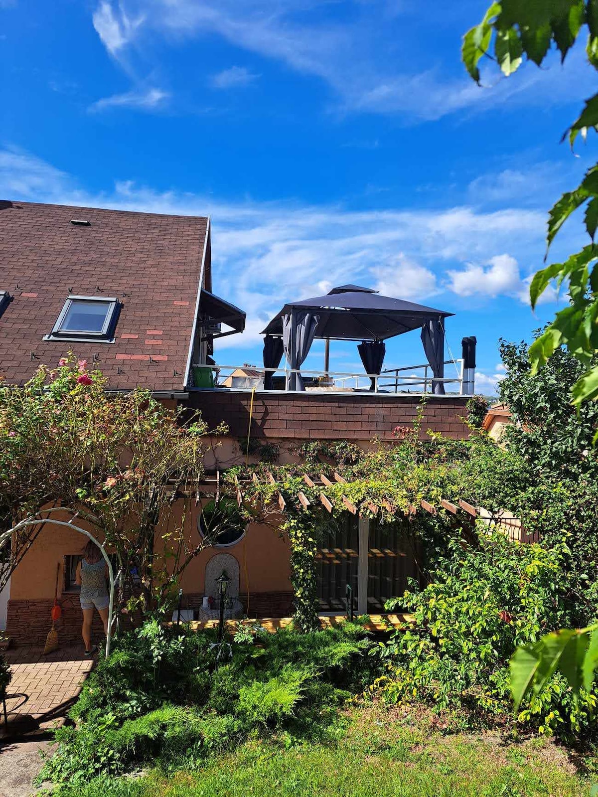 A two-story structure displays a designed rooftop terrace with a shaded area, featuring a canopy and outdoor seating. Lush greenery surrounds the building, with flowers and shrubs framing the image. The sky above is clear with scattered clouds, enhancing the tranquil outdoor setting.