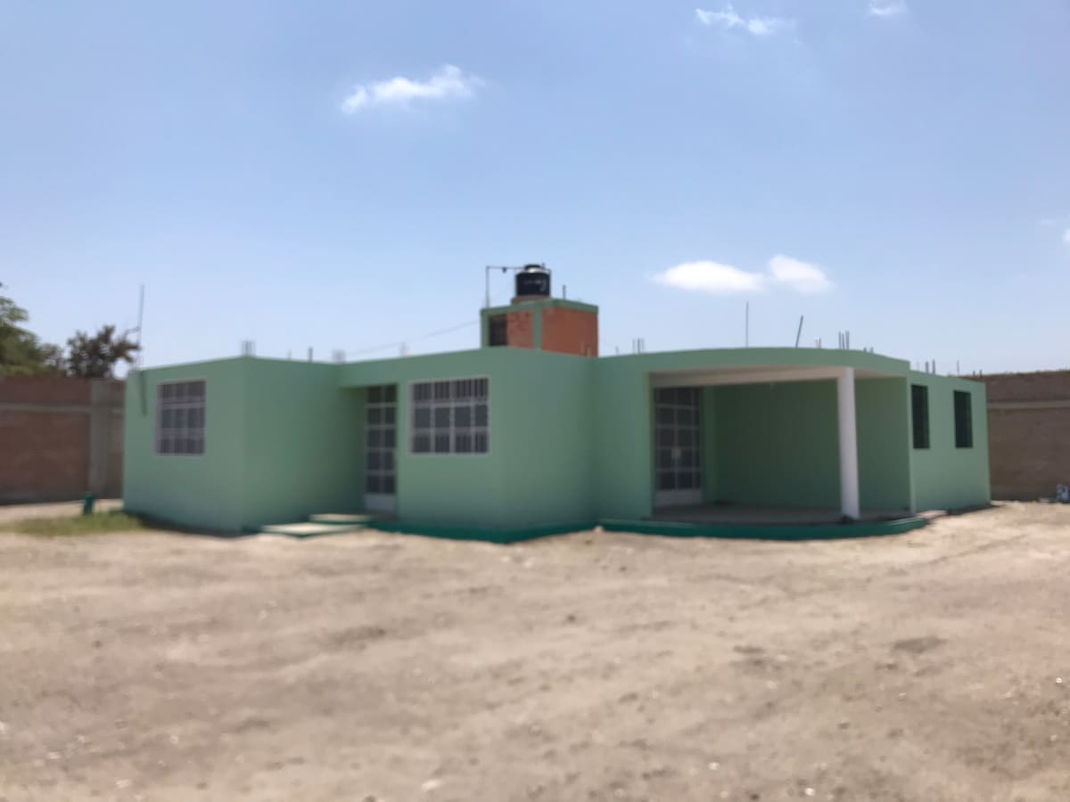 The exterior of a spacious green building is displayed, featuring multiple large windows that allow natural light. A covered porch extends from the entrance, while the surrounding area is open with gravel underfoot, providing ample space for activities.