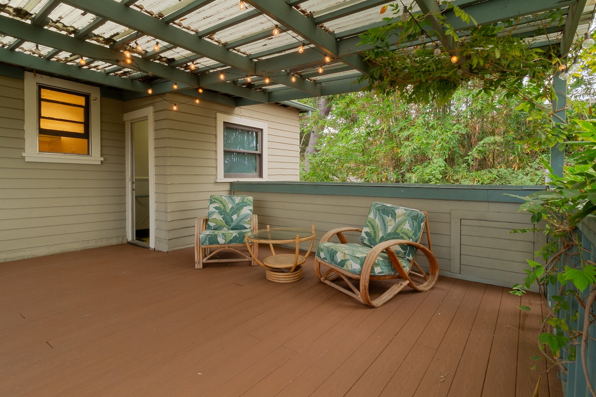 A spacious outdoor deck features wooden flooring and overhead string lights. Two comfortable chairs with tropical patterns are positioned on the deck, accompanied by a natural wood side table. Lush greenery is visible in the background, providing a relaxing atmosphere for alfresco activities.