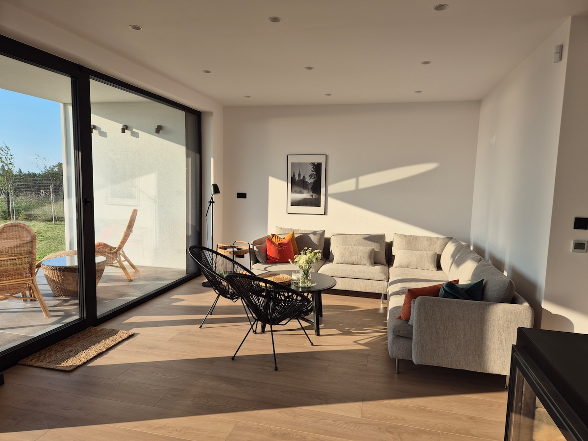 A modern living area is displayed with large glass doors inviting natural light. A sectional sofa and lounge chairs are arranged around a coffee table. Decorative cushions and a small vase with flowers are positioned on the table, enhancing a comfortable ambiance.