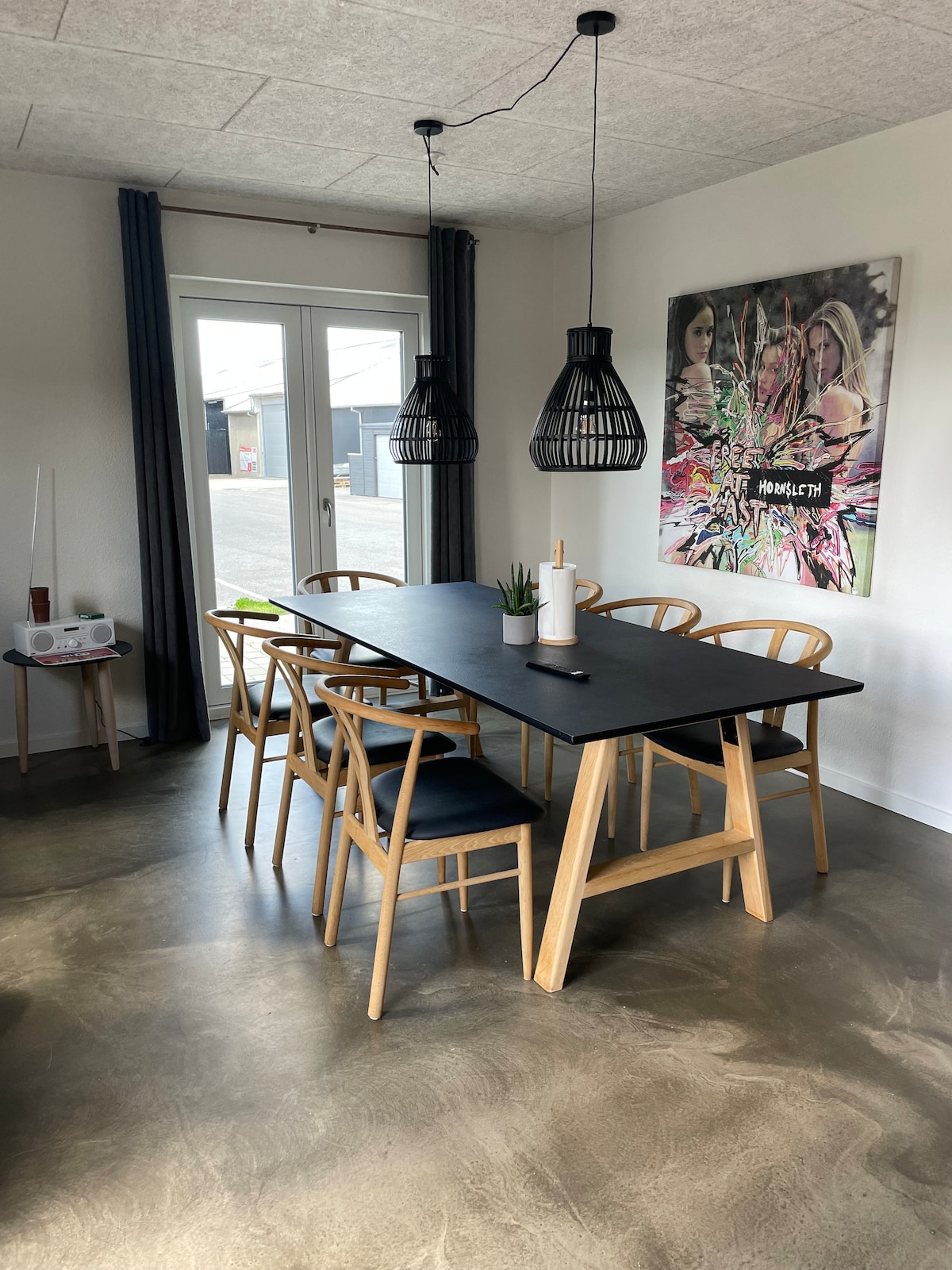 A dining area features a long black table surrounded by six wooden chairs. Two pendant lights hang above the table, illuminating the space. A large piece of wall art is displayed nearby, and large windows allow natural light to enter, with drapes partially covering them.
